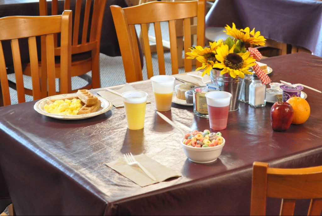 A table with a maroon table cloth and wooden chairs around it on the table are sunflowers as a centerpiece and breakfast set out