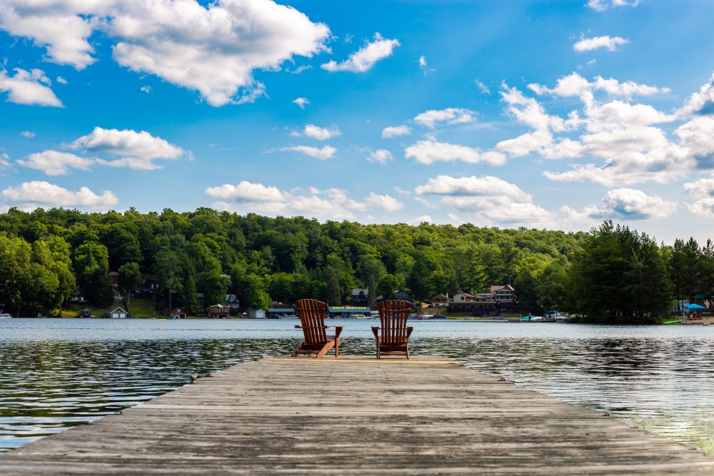 A dock facing the clam lake and green trees on the other side on the dock there are two Adirondack chairs