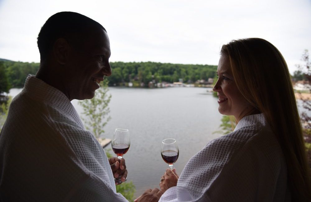 A couple in bathrobes on a porch sipping wine with the lake in the background