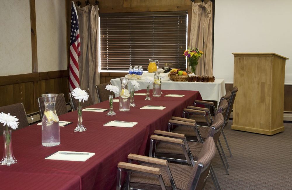 A long table with a red table cloth and chairs set up around it on the table are single white flower centerpieces and pitchers of lemon water and the end of the table is a buffet table with breakfast items on it and next to the buffet is a wooden podium