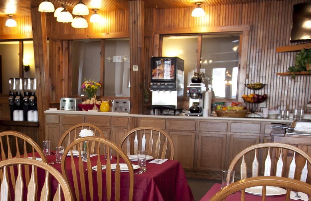 A restaurant with maroon table cloths a centerpiece of a large white flower, wooden chairs and place settings set at each table