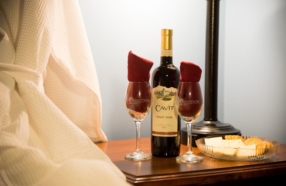 A bedside table with a plate of cheese and crackers and a bottle of wine as well as two wine glasses with napkins nicely placed in the glasses