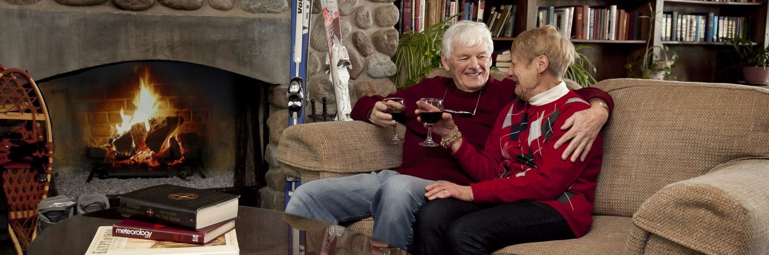 An old couple sitting together on a couch by a roaring fireplace, smiling at each other with wine glasses in their hands