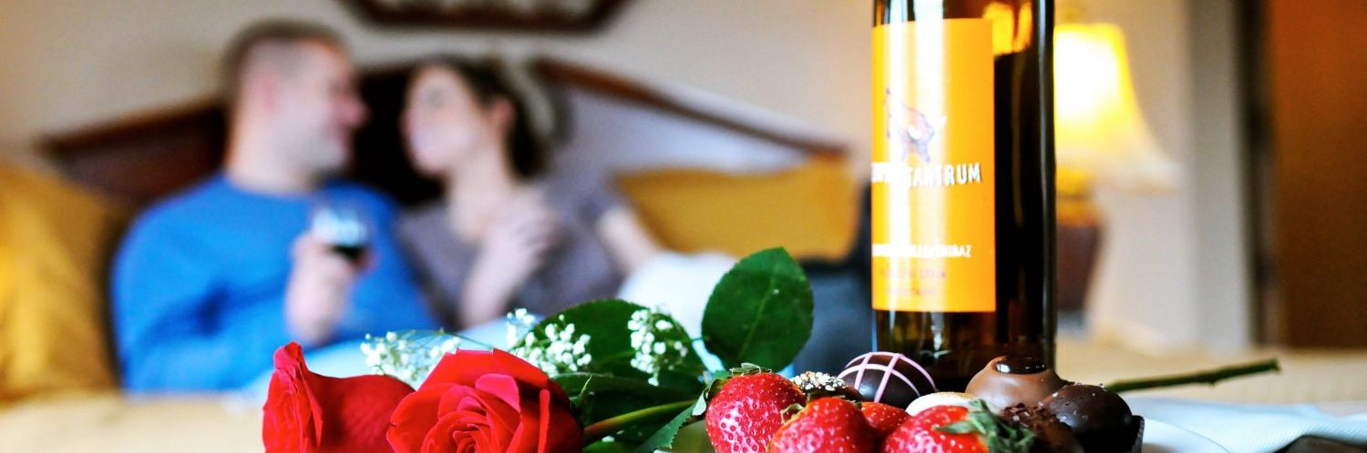 An image of a couple smiling at each other sitting on a bed with wine glasses in their hands in the forefront of the photo there are two roses, a plate oof strawberries and chocolate and a bottle of wine