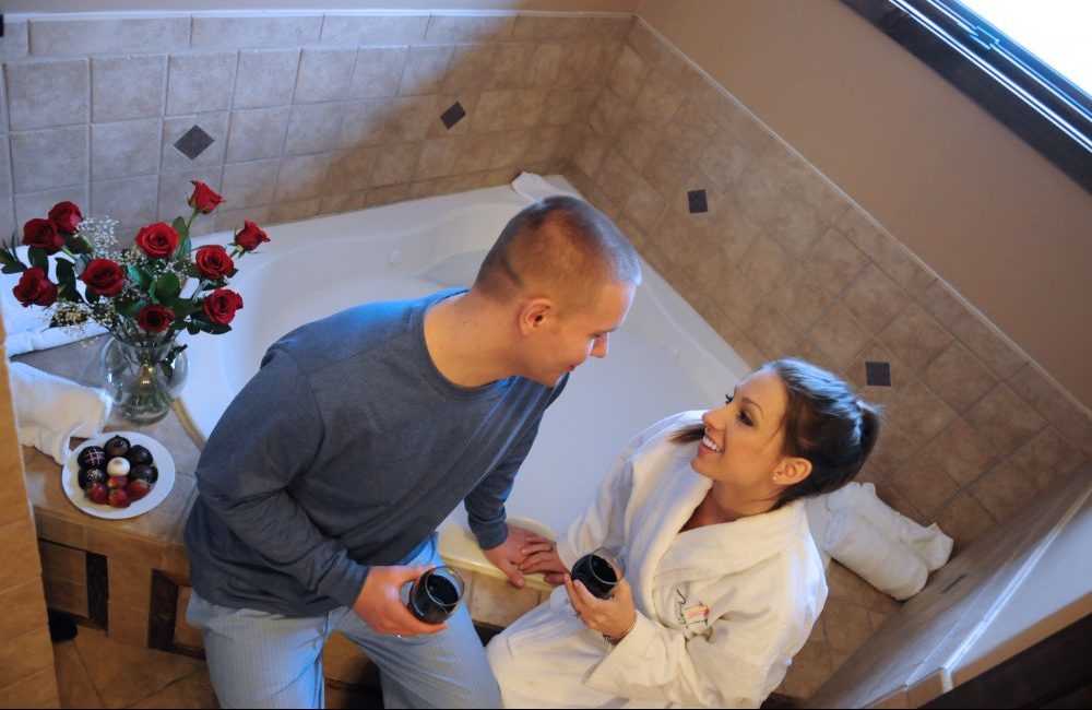 A couple sitting on the side of the bathtub drinking wine and smiling next to them is a plate of chocolates and strawberries and a vase of roses