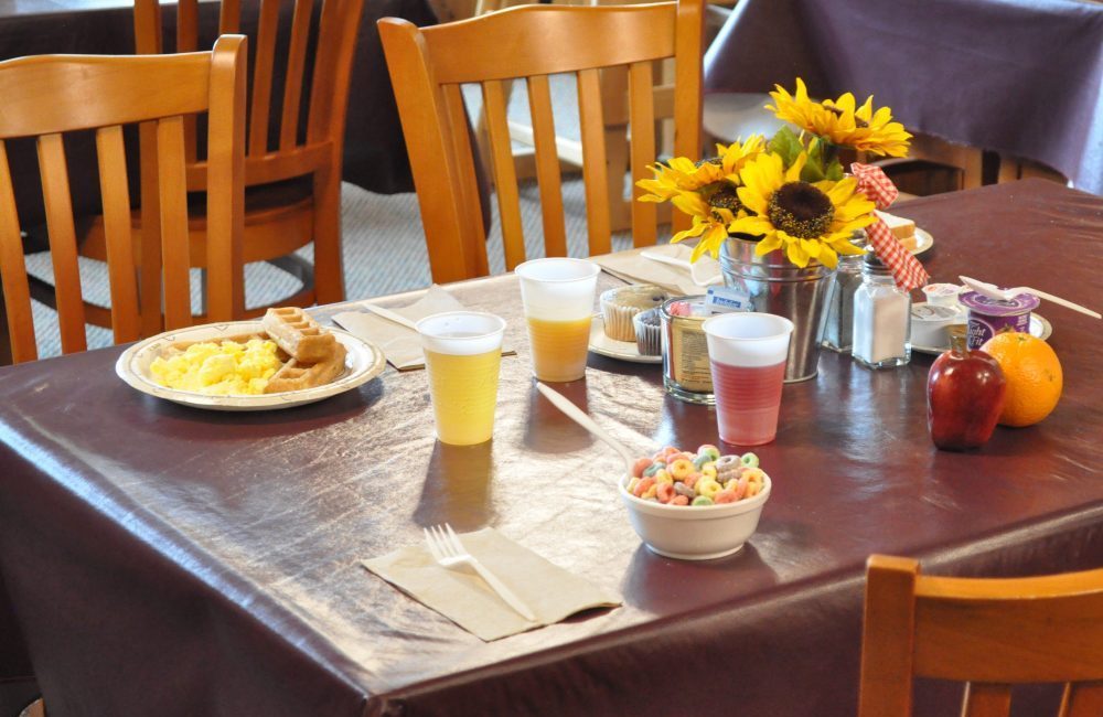 A table with a maroon table cloth and wooden chairs around it on the table are sunflowers as a centerpiece and breakfast set out