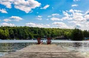 A dock facing the clam lake and green trees on the other side on the dock there are two Adirondack chairs
