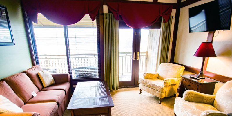 A living room with large windows and French doors that lead to a porch in the living room there is a couch, coffee table, two chairs, a stand with a lamp on it, and a flat screen tv mounted on the wall