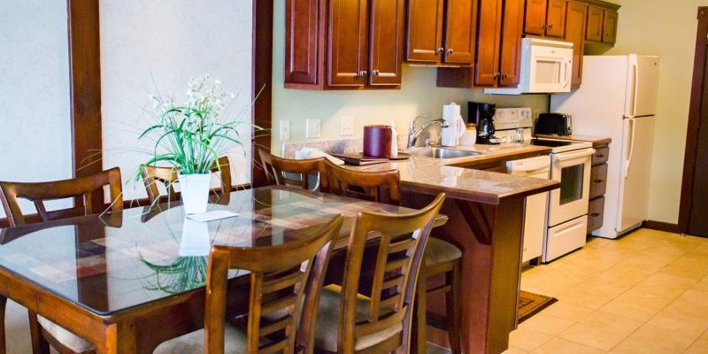 A kitchen with wooden cabinets, a stove/oven, a coffee maker, dishwasher, fridge, and a microwave there is also a wooden table with wooden chairs with cushions