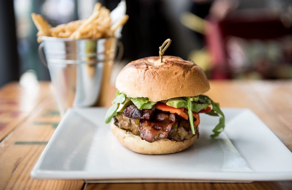 A bacon burger on a white plate with a bucket of fries