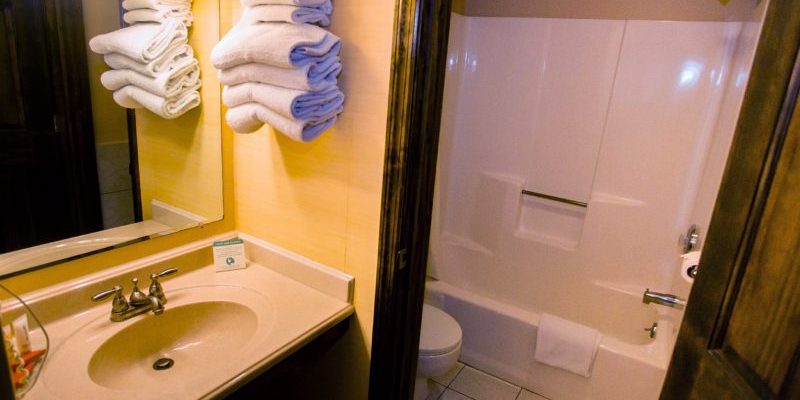 A white toilet and shower and a door that separates them from the sink and towels folded and stacked on a shelf next to the sink