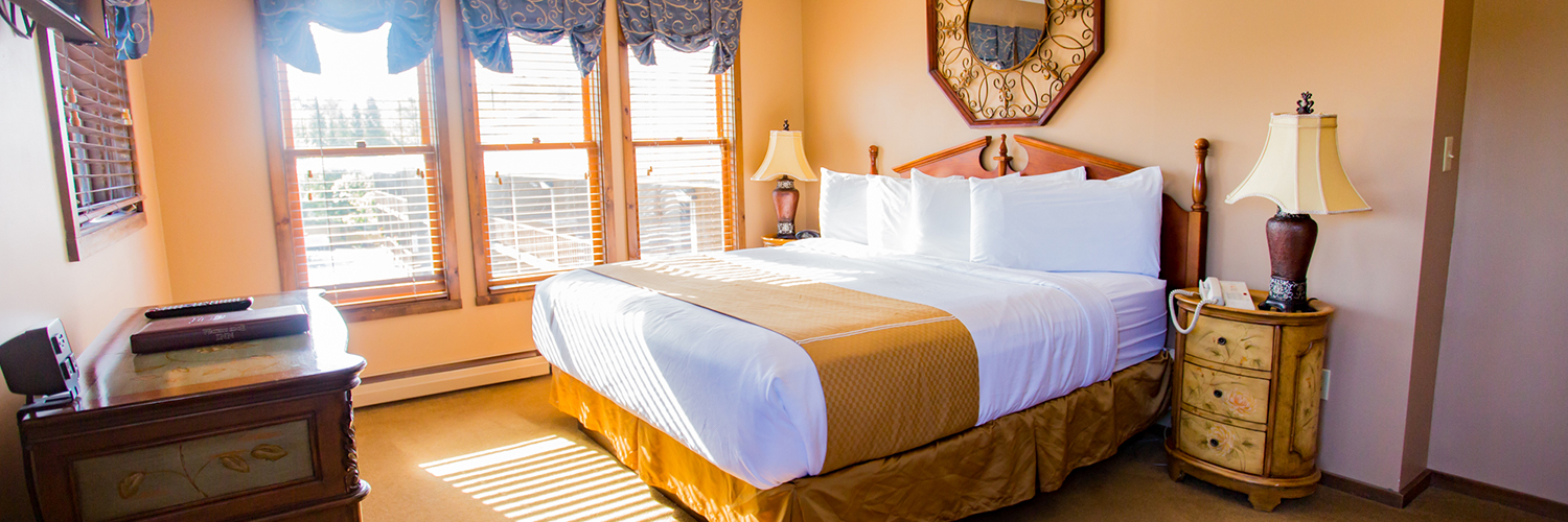 A hotel room with a large bed, two nightstands, a dresser, a tv hung on the wall, and 4 windows letting a lot of natural light into the room