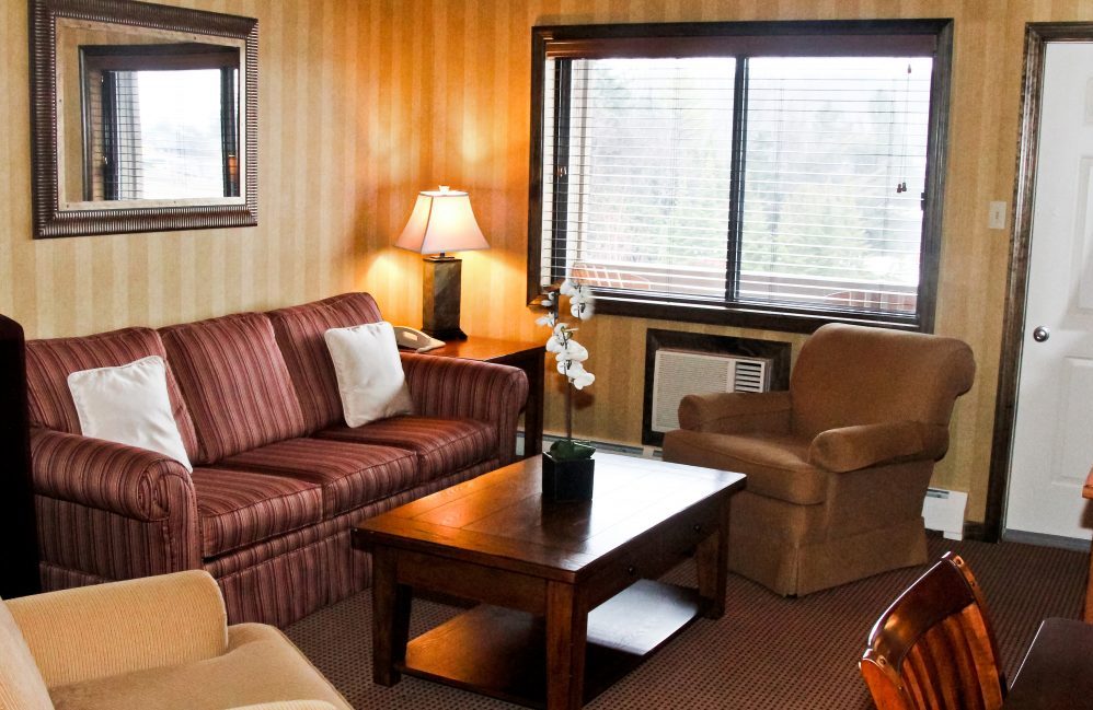 A living area with a couch a coffee table with flowers on it, a stand with a lamp on it, a comfy chair, and a desk