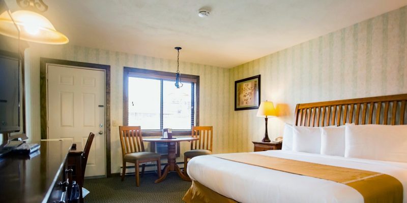 A hotel room with a king size bed as well as a dresser with a flat screen tv and to the side of the room is a window with a small table and two chairs set up beneath it