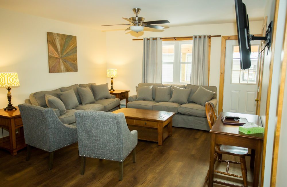 A living room with two couches, two chairs, wooden table, two lamp tables with lamps, a desk, and a tv hanging on the wall