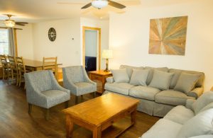 A living room with two couches, two chairs, wooden table, two lamp tables with lamps, and behind the living room is a dining room table