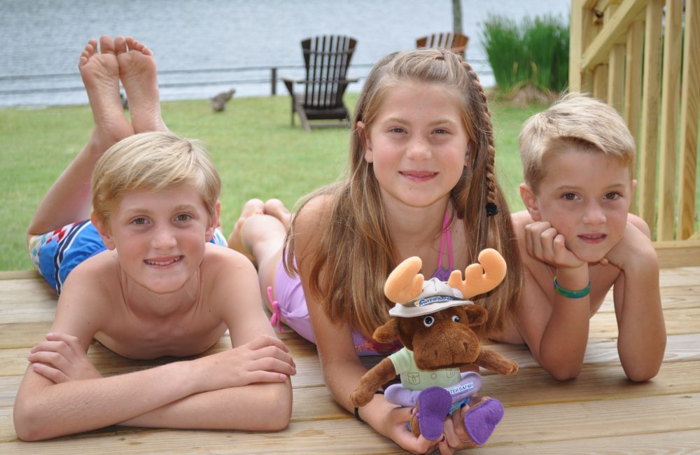 Three kids on a porch in front of the lake in their bath suits and a stuffed Walter Safari