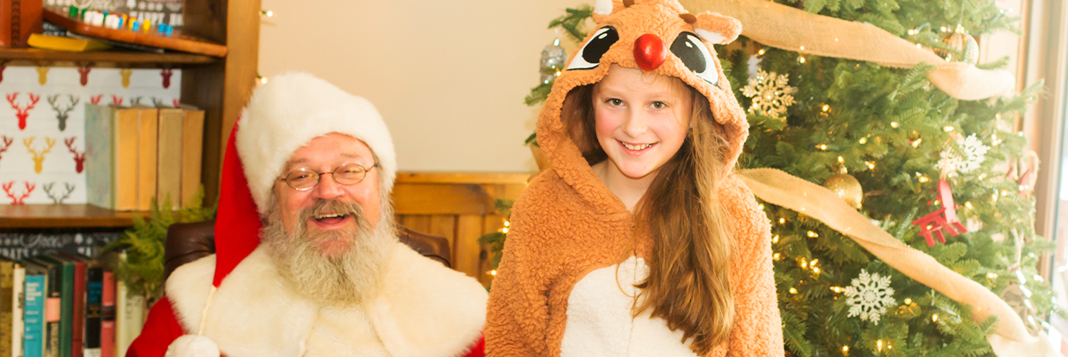 a girl in a Rudolph costume sitting on Santas lap next to a Christmas tree